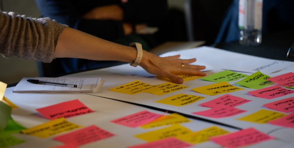 Mano di una partecipante che organizza post-it colorati su grandi fogli durante un laboratorio di progettazione partecipata.