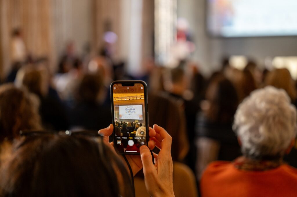 Una persona tra il pubblico fotografa con lo smartphone il palco di un convegno; sullo schermo del telefono compare la scritta “Ponti di Cultura”, mentre la sala affollata resta sfocata sullo sfondo.