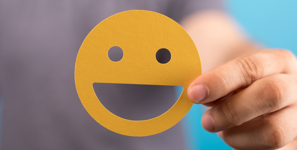 Mano che regge una sagoma gialla con un volto sorridente, simbolo di benessere, soddisfazione e valutazione positiva.